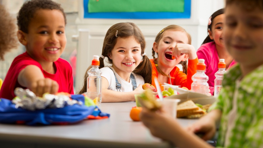 5 young children sit around a cafeteria table eating school lunch